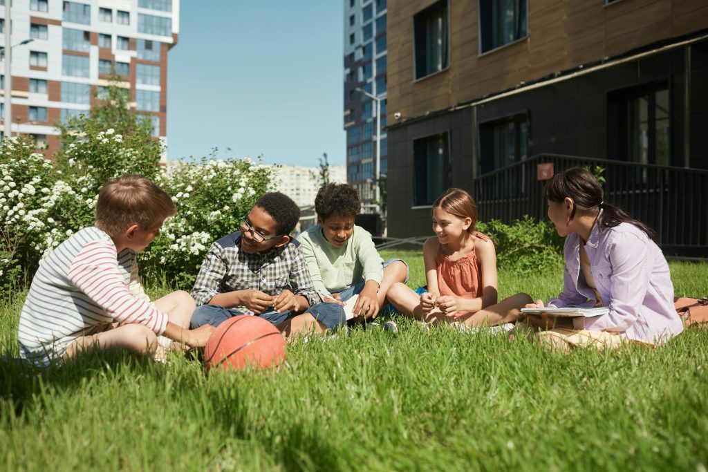 pexels-photo-8457811-8457811 A diverse group of kids sitting on grass, having fun together under a clear blue sky.
